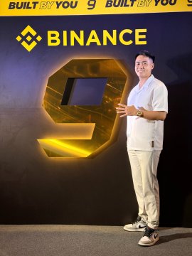 A photo of a man in a white shirt standing in front of a large yellow '9' logo and the word 'BINANCE' with the slogan 'BUILT BY YOU'.