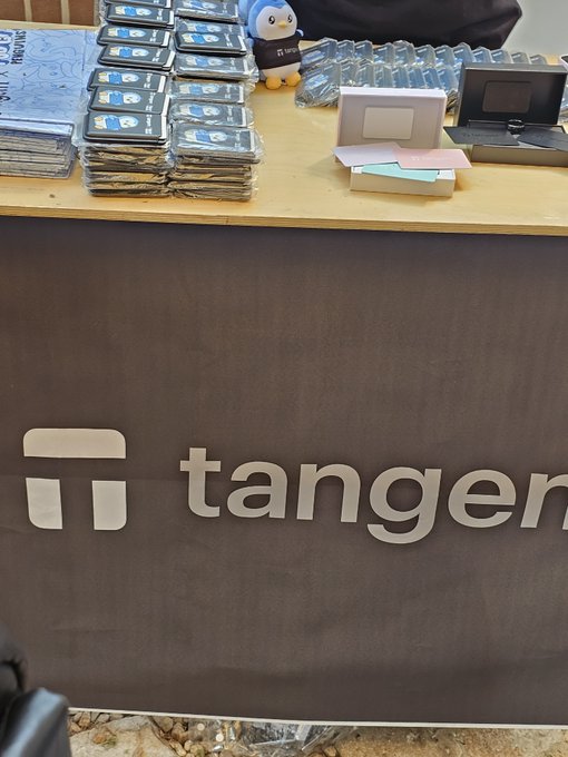 A booth displaying the 'tanger' logo on a dark banner, with stacks of packaged products, a plush mascot figure, and water bottles arranged on the counter.
