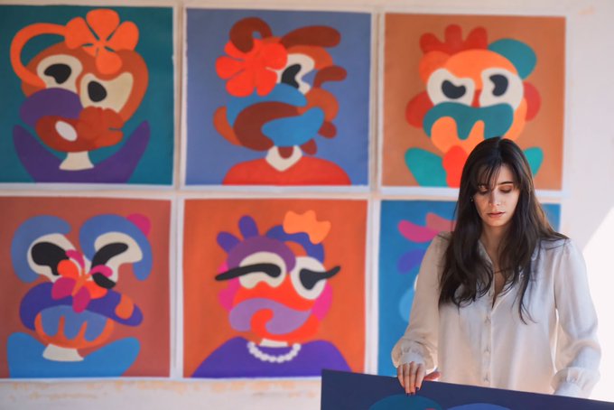 A woman in a light-colored blouse stands in front of six colorful, abstract portraits featuring stylized faces with bold, graphic shapes.