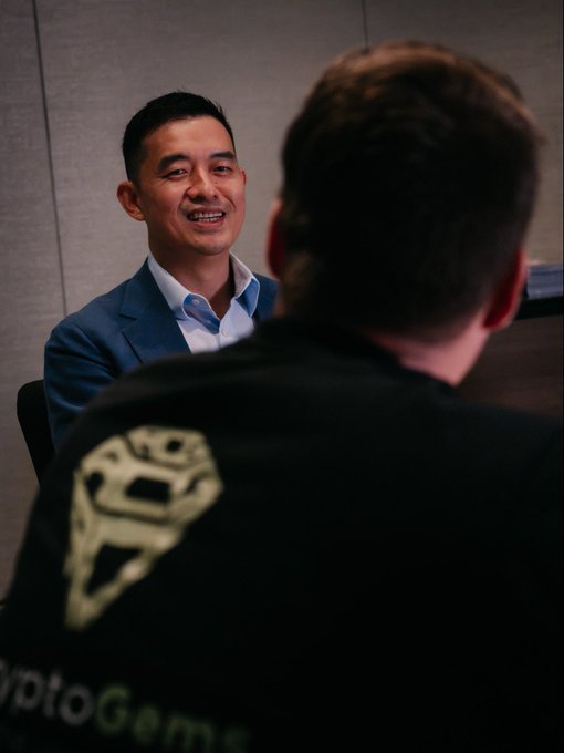A man in a blue suit smiles warmly during an interview with someone wearing a "CryptoGame" branded black shirt, capturing a moment from the conversation with Han, founder of Gate, as they discuss his 13-year journey building the exchange.