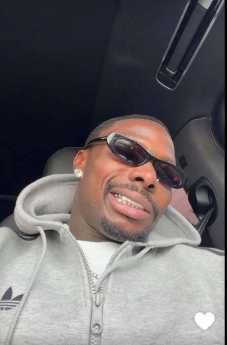 A selfie of a man wearing dark sunglasses, a diamond earring, and a gray Adidas zip-up hoodie, sitting inside a car with a white heart icon visible in the corner.