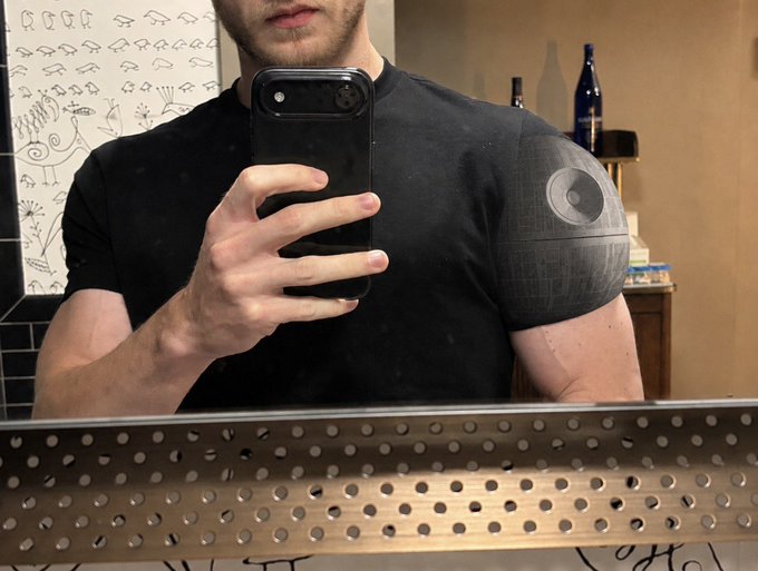 A man takes a mirror selfie in a bathroom, holding up a smartphone to showcase a detailed Death Star tattoo on his left shoulder that seamlessly blends into the black t-shirt's dark fabric. The iconic spherical space station artwork on his deltoid matches the "Death Star Delts" caption, cleverly creating the illusion that the tattoo is a permanent fixture of his shirt.