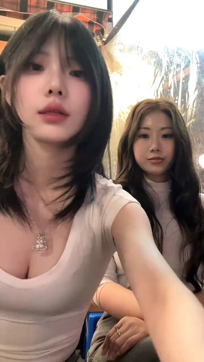 The image features two women with long hair, both wearing white tops. They are standing close together, possibly posing for a photo or video. The background of the image appears to be a gray wall, which contrasts with their attire. There is also an element in the image that seems unrelated to the main focus - a post on social media. This post contains text about Bitcoin and Etherium, suggesting that it might be related to cryptocurrency or digital currency discussions.