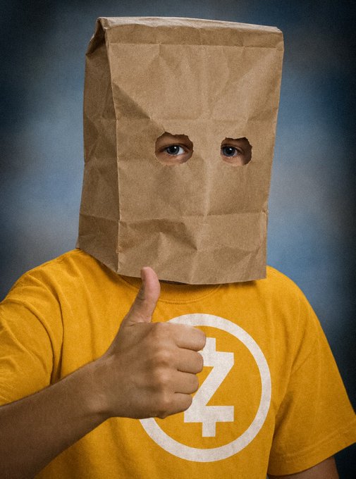 A person wearing a yellow Zcash (ZEC) t-shirt gives a thumbs-up while wearing a brown paper bag over their head with holes cut out for their eyes, humorously relating to the $ZEC and $ZCAT cryptocurrency tickers by depicting anonymous approval.