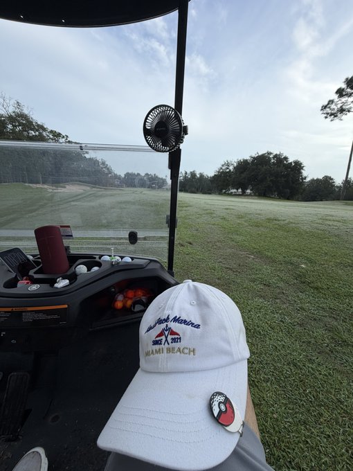 A view from a golf cart shows a white cap with "Black Mesquite 2017 MIAMI BEACH" text and a ball marker resting on someone's lap, with golf balls and a fan visible on the cart and a green course in the background.