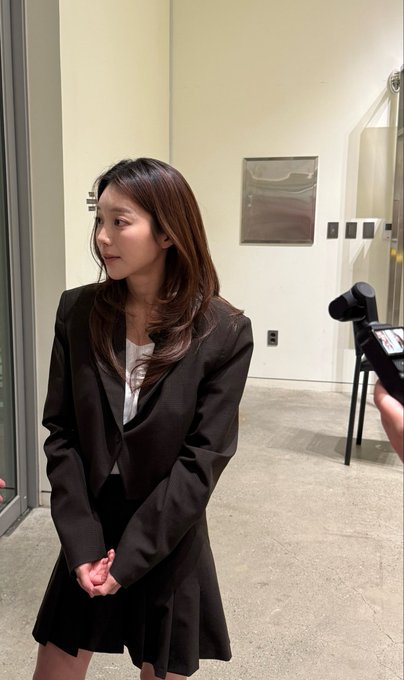 A young woman in a brown blazer and skirt stands with her hands clasped politely in front of her, looking to the side with a soft expression, while a camcorder is visible on the right being held toward her. The image visually supports the caption's description of "JADOODOO" being "genuinely so gentle offline," capturing her poised and shy demeanor as she appears to be filmed during an interview or media interaction.