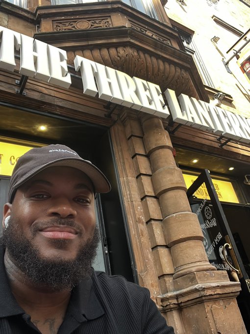 A selfie of a man wearing a black cap and shirt with AirPods, smiling in front of a classic pub with a sign reading 'THE THREE LANTERNS'.