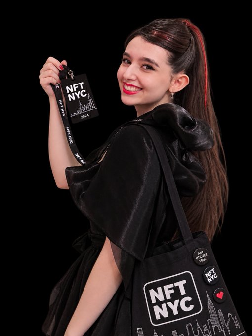 A smiling woman with a ponytail holds an 'NFT NYC 2024' badge and a tote bag against a black background, wearing a black hooded outfit.