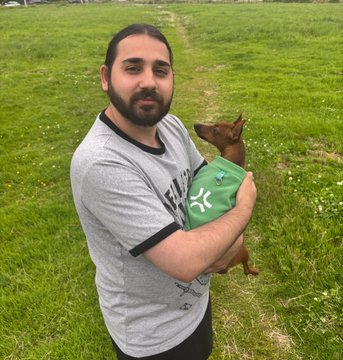 A man with a beard in a gray t-shirt stands in a grassy field holding a brown dog wearing a green harness.