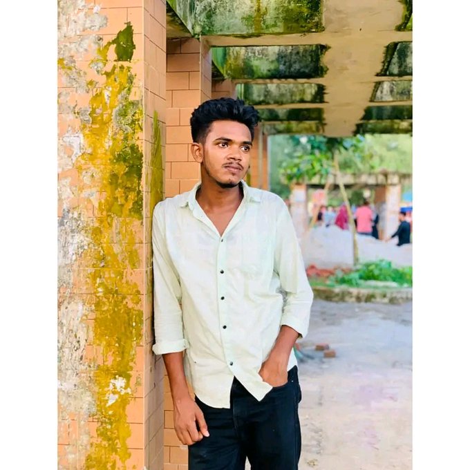 A young man in a light green shirt leaning against a weathered, yellow-painted brick wall outdoors.