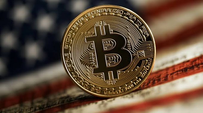 A physical Bitcoin token standing upright against a blurred background of the American flag.