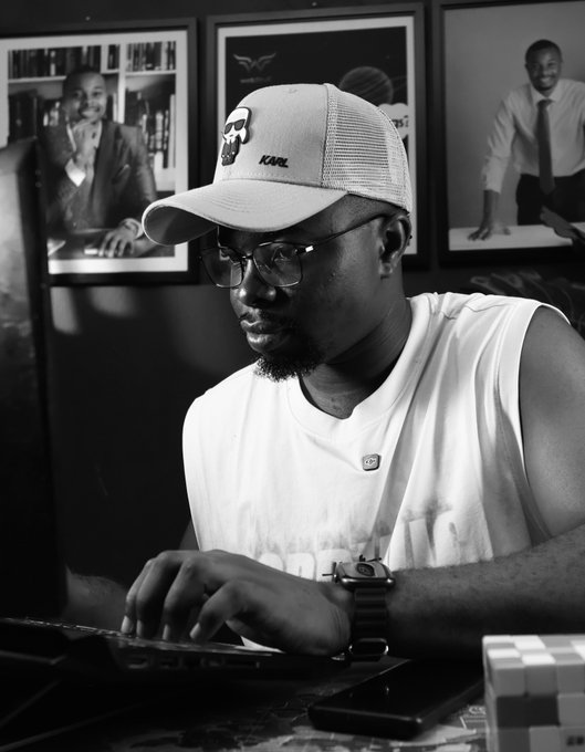 A black and white photo shows a man wearing a trucker hat, glasses, and white tank top working intently at a laptop, with framed photos visible on the wall behind him.