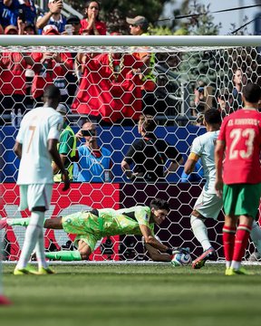Action shot from a soccer match showing a goalkeeper diving to make a save while players from opposing teams, including one wearing a #7 white jersey and another in a red #23 jersey (A. Hakimi), look on.