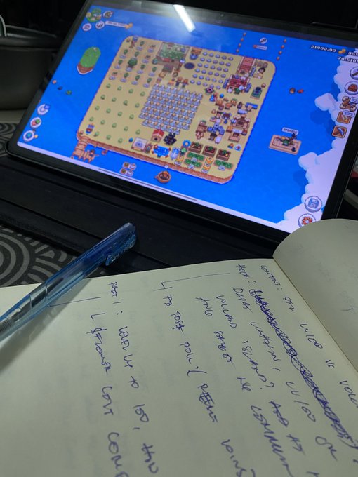 A tablet displaying a farm/island-building game next to an open notebook with handwritten notes and a blue pen.