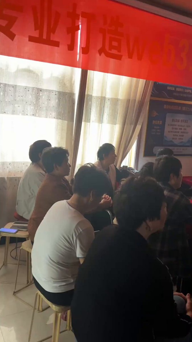The image shows a group of people sitting around a table with chairs, engaged in conversation or possibly attending an event. The room has red curtains that add color to the scene. There are at least nine visible individuals in the image, some of whom appear to be wearing ties. A cell phone can also be seen on one of the tables.

The post features a red banner with Chinese characters and English text, which reads "I think, haha, the most successful CX project in recent years (negative connotation— Pi Coin). I've already seen a ton of these grassroots promotional videos, all featuring uncles and aunties, not even a single middle-aged person. After Pi Coin launches, whether this group actually". The image seems to be related to the post's content or message about the Pi Coin project.