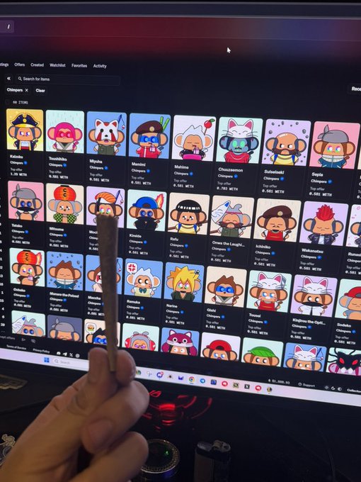 A hand holding what appears to be a joint or blunt in front of a computer monitor displaying a marketplace of cartoonish monkey NFT characters.