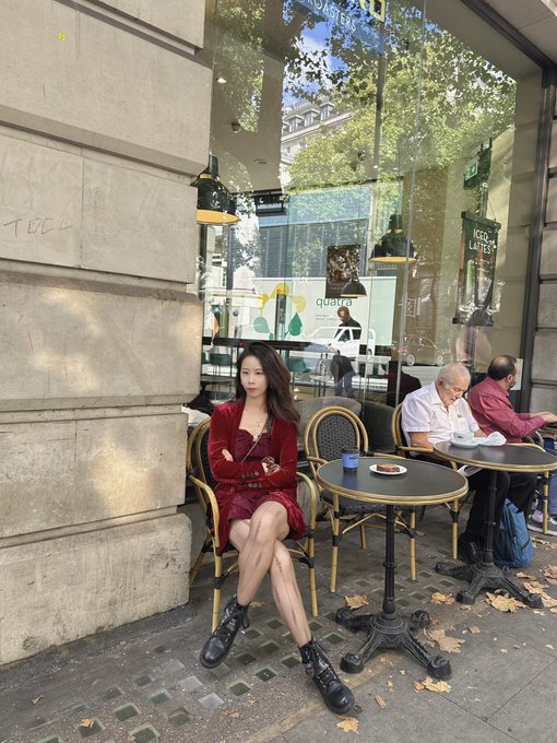 A young woman in a red outfit sits at a classic Parisian cafe terrace, with an older man in a white shirt seated at the next table reading papers.