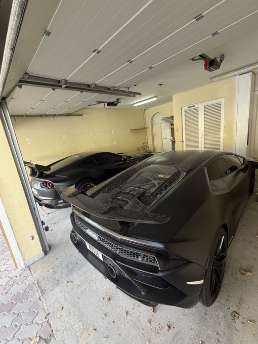 Two matte black sports cars—a Chevrolet Corvette and a Toyota GR Supra—are parked side by side inside an open garage. The post "GM" likely refers to General Motors, tying the Corvette to the GM brand, while the image appears to showcase two high-performance cars in a collector's garage.