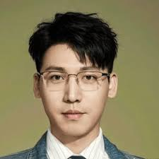 A professional headshot of a young East Asian man wearing rimless glasses, a striped shirt, tie, and denim jacket against a plain greenish-yellow background.