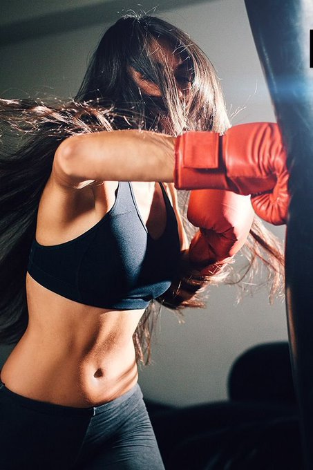 A woman in athletic wear and red boxing gloves throwing a punch at a punching bag in a dramatically lit setting.
