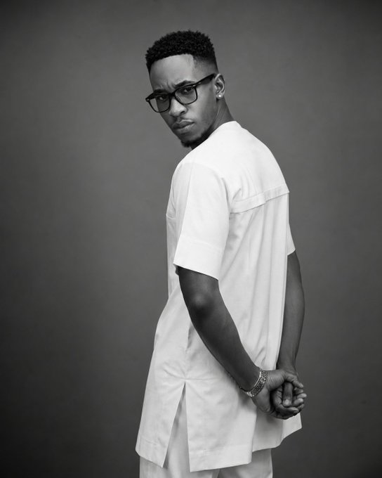 A black-and-white portrait of a man with short hair, glasses, and a short beard, wearing a white layered outfit, photographed from the side against a gray background.