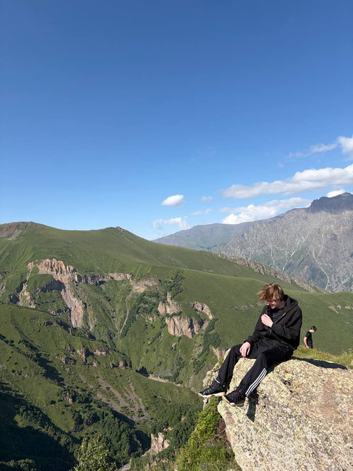 A person in a black tracksuit sits on a rocky outcrop overlooking a vast mountain range with green slopes and a deep valley under a blue sky.