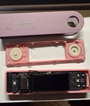 A disassembled pink Ledger hardware wallet reveals its internal components, including the circuit board and screen assembly, with the device's casing opened to expose potential tampering areas. The image illustrates claims of a hidden SIM card implant allegedly discovered inside the wallet's display padding, linked to the reseller "CryptoBilis."
