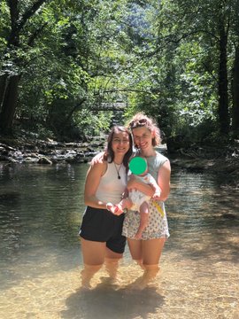 Two young women stand in a shallow river surrounded by a lush forest, with a green emoji covering one woman's face.