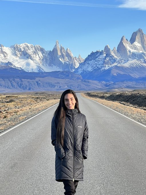 A woman with long dark hair wearing a black puffy coat standing on an empty road with the snow-capped peaks of Mount Fitz Roy in the background.