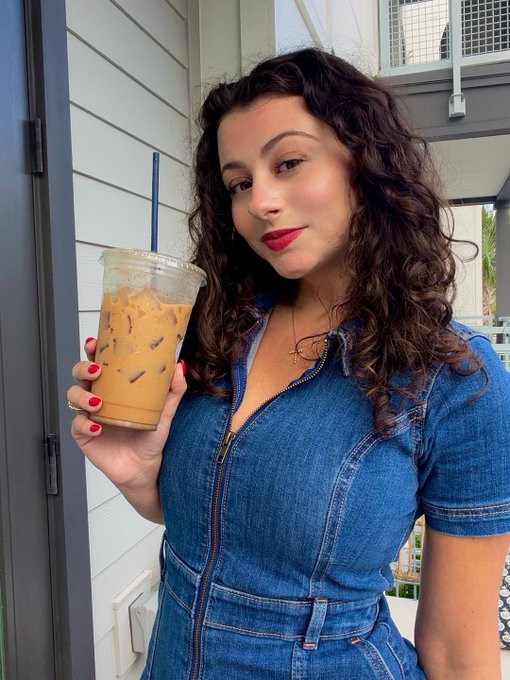 A woman with curly dark hair, red lipstick, and a denim zip-up dress holds up an iced coffee in a clear plastic cup while standing outside near a building with white siding.