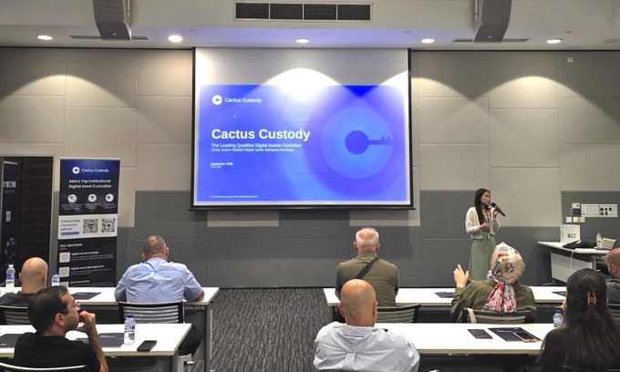 A woman presents in front of a large screen displaying "Cactus Custody - The Leading Qualified Digital Assets Custodian" while addressing an audience at what appears to be a conference or seminar setting, with attendees seated at tables facing the presentation. A roll-up banner beside her advertises Cactus Custody's institutional digital asset services, supporting the context of the post about hosting a Turkish Automotive Leadership Delegation to discuss stablecoin custody solutions for dealers.