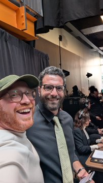 A selfie taken in what appears to be a podcast or studio setting shows a smiling bearded man in a green cap and glasses alongside another smiling man in a black shirt and tie.