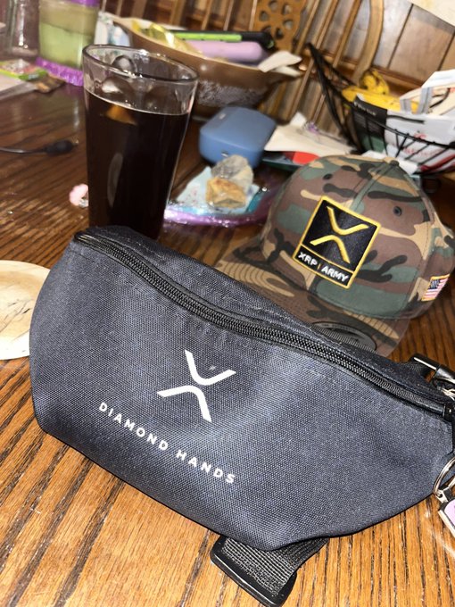 Casual photograph of a table showing a glass of iced dark drink, a camo XRP Army cap, and a black bag with the XRP logo and 'DIAMOND HANDS' printed on it.