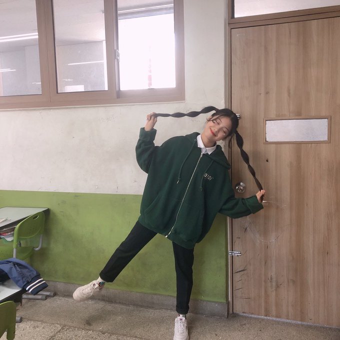 A young woman wearing an oversized dark green hoodie and black pants playfully poses by holding her long braided pigtails in what appears to be a classroom setting.