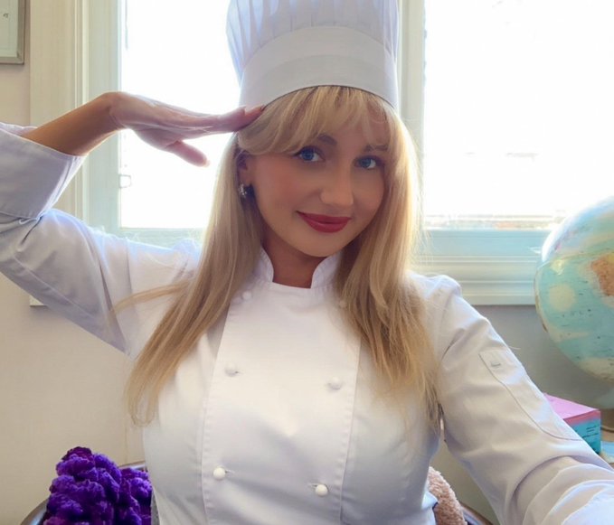 A blonde woman wearing a white chef's hat and chef's jacket, saluting with her right hand near her forehead.
