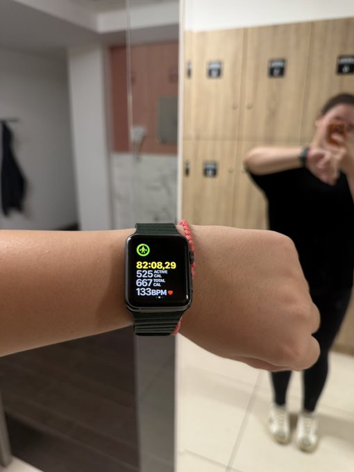 A person holds up their wrist showing a smartwatch displaying a workout summary at a gym locker room, with stats including a time of 02:08:29, 925 CAL active, 657 CAL total, and 133 BPM heart rate, while their reflection is visible in the mirror.