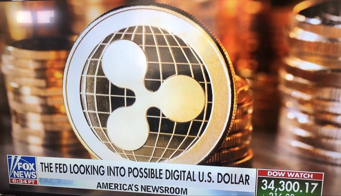 A close-up of a gold XRP coin sits atop stacks of other cryptocurrency coins, with a Fox News chyron reading "THE FED LOOKING INTO POSSIBLE DIGITAL U.S. DOLLAR" and a Dow Jones indicator showing "34,300.17" in the background—tying into the post's claim about XRP's potential global financial dominance.