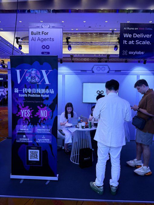 The image features a group of people standing inside an exhibition hall, looking around at various booths set up in front of them. One of the booths appears to be related to artificial intelligence or machine learning projects, as indicated by its name tag. There is also a sign near the booth that reads "Built for AI agents" and another one that says "Vox". People are standing next to a table displaying some laptops. They seem to be interested in exploring different exhibits at the venue.