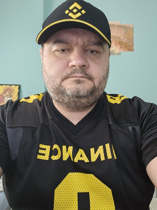 A man wears a black and yellow Binance jersey with the text "BINANCE" and number "0" visible, paired with a matching Binance cap featuring the logo. The branded outfit aligns perfectly with the "Binance Vibes" caption, showcasing his enthusiasm for the cryptocurrency exchange.
