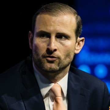 A close-up shows Patrick Witt, the White House's crypto policy advisor, wearing a navy suit and peach-colored tie, appearing to speak at an event with a dark blue-lit background. His serious expression aligns with the urgent tone of the accompanying post, which reports his warning that Congress is running out of time to pass the Crypto Clarity Act amid disagreements over provisions limiting President Trump's crypto profits.