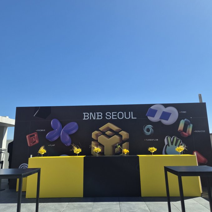 An outdoor promotional display for 'BNB SEOUL' features a black backdrop decorated with colorful 3D logos including PRIMEUS, FLAP, COMBO, RENATA, and the BNB Chain emblem, with yellow and black counter displays and flower arrangements in the foreground.