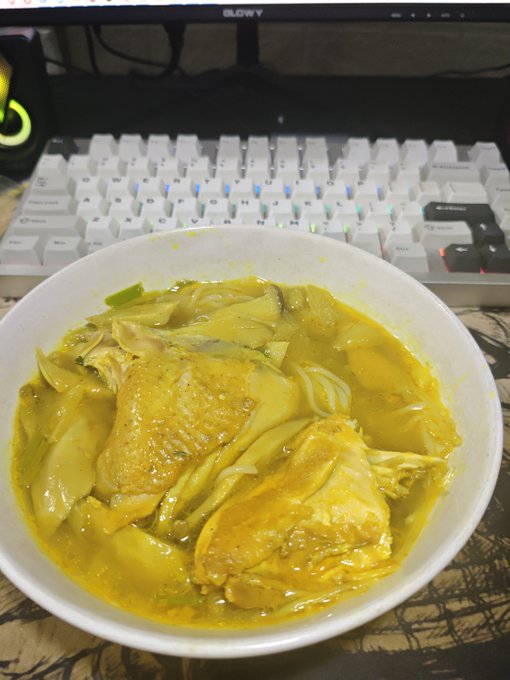 A bowl of yellow chicken curry with bamboo shoots and basil leaves, placed on a desk in front of a white glowing computer keyboard and monitor.