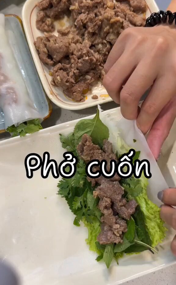 The image shows a person preparing food, specifically making a roll with meat and greens. The person's hands are seen cutting up the ingredients for the roll while wearing a black bracelet. There are also other items present in the scene, such as a bowl of food on a table and another plate nearby. The post appears to be about cooking or sharing recipes, possibly related to Asian cuisine.