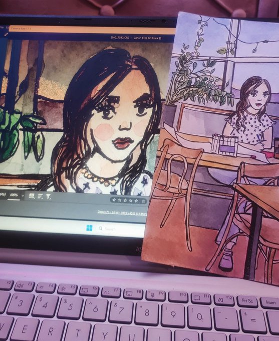 A laptop screen displaying an illustration of a girl with large eyes, with a physical watercolor print of a similar girl sitting at a café table placed on the keyboard.
