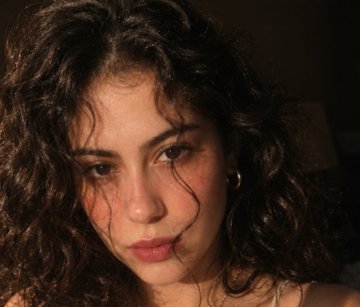 A close-up portrait of a young woman with curly brown hair and freckles, lit with warm light against a dark background.