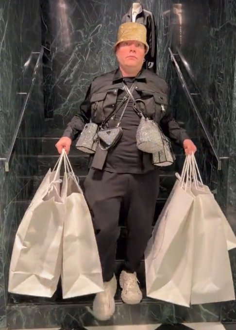 A person wearing a gold sequined bucket hat and a black outfit stands on marble stairs holding multiple large silver shopping bags and wearing sparkling silver purses.