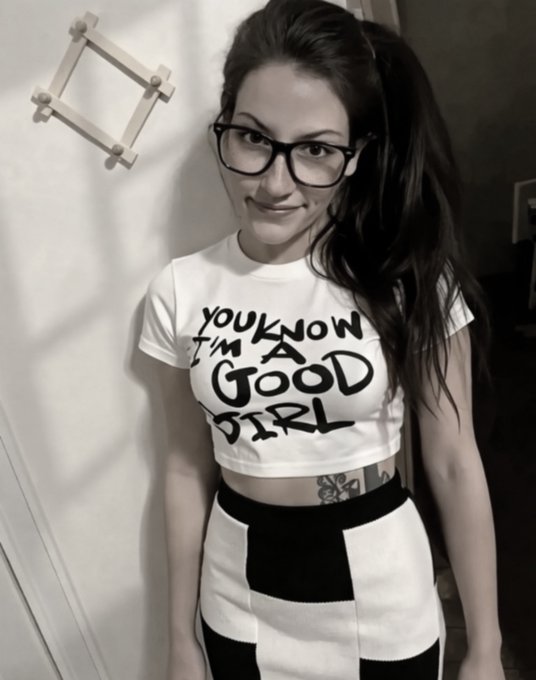 A black and white photo of a woman wearing glasses and a white cropped t-shirt that says 'YOU KNOW I'M A GOOD GIRL', paired with a black and white checkered skirt.