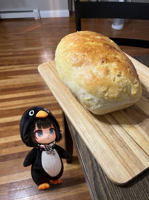 A small penguin-themed chibi plush doll stands next to a freshly baked loaf of bread on a wooden cutting board.