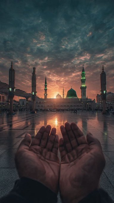 A first-person view of two open hands reaching toward a mosque with green domes at sunset.