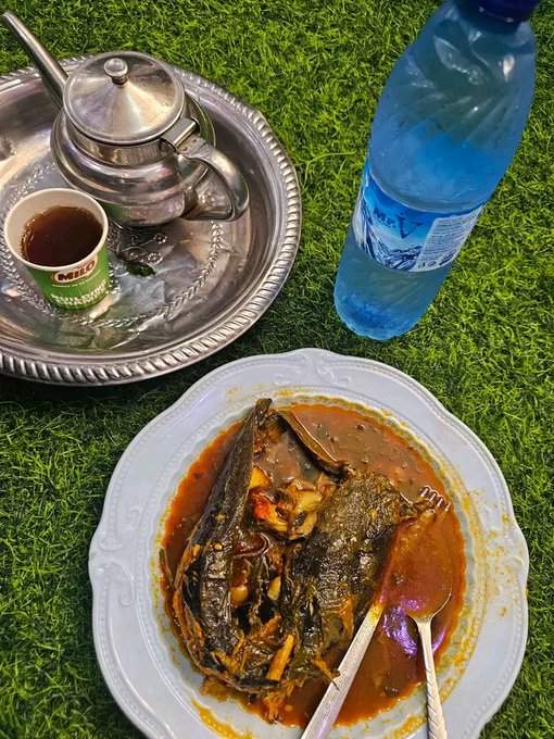 A traditional Middle Eastern-style cooked fish head dish served in red sauce on a white plate, alongside a silver teapot with tea in a small cup and a blue bottled water, all set on artificial green grass.